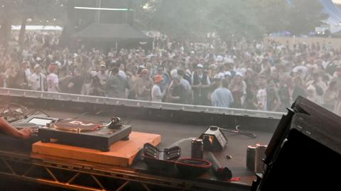 A set of DJ decks in the foreground with a crowd of people in the background, at a music festival. There's trees in the background behind the crowd. The crowd themselves are dancing, all are wearing various items of summer gear like shorts, T-shirts and bucket hats.