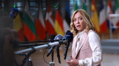 Giorgia Meloni stands in front of a cluster of microphones, wearing a white shirt and a pink waistcoat. 