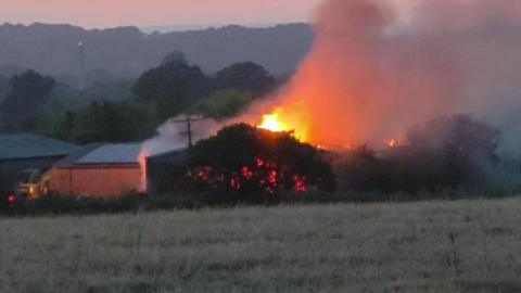 A zoomed in picture showing the farm fire from a distance. It appears to be affecting a large wooden barn across the field, hidden behind a bush. There are bright orange flames bursting from the roof and smoke billowing across to the right. It is dusk and the sky is a faded orange.