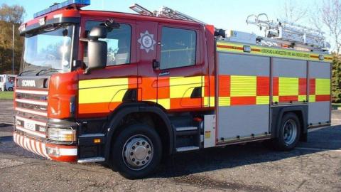 A fire engine pictured from the side. It is a red, yellow and grey truck with the words Hereford and Worcester Fire and Rescue Service on the side.