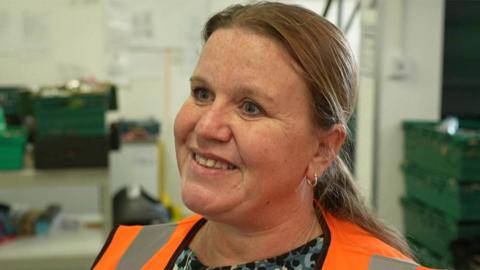 Close up of Lisa Parkinson who stands in the warehouse in front of green food crates. She is wearing an orange high vis vest and has long fair hair tied back in a ponytail
