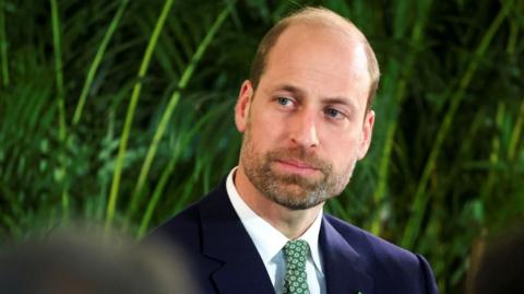 Prince William wearing a dark suit with a green tie against a backdrop of greenery