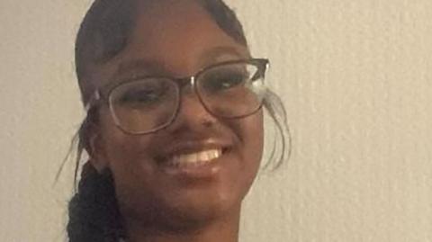 A close-up photo of a smiling Elianne Andam, she has long black hair in a braid and is wearing glasses. She is wearing a striped school uniform blouse.