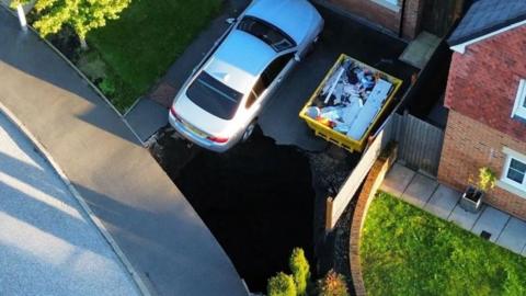 Drone shot of the massive black hole on the driveway of a new build house. There is a silver car between the hole and the front of the house with the back of it hanging over the hole and a full skip to the side of the car