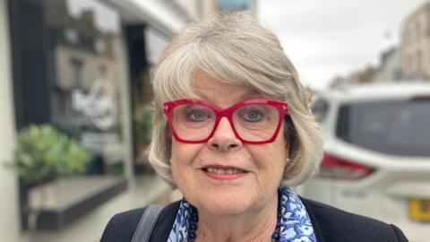 A woman with red glasses looks directly at the camera, she has grey bobbed hair. She is wearing a jacket and a blue patterned shirt. She has a handbag strap over her shoulder and is wearing a necklace and earrings.