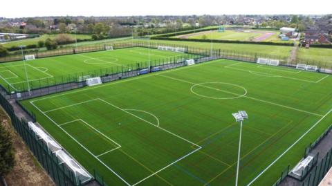 Two football pitches side by side, with markings and a secure fence around. Both have floodlights and a running track is in the distance.