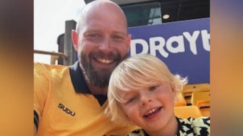 Tom, who has a beard, is wearing a Wolves shirt and smiling next to son Rory, who has blond hair. They are both looking at the camera and seats at Molineux are visible behind them.
