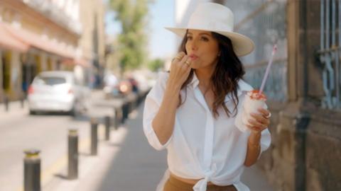 Eva Longoria wearing a white hat and shirt walking down the street eating an ice cream