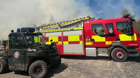 A small black vehicle with the fire service logo on the side. On the back of its trailer it has yellow pipes. Next to it is a red and yellow fire engine with a metal grey ladder on top. There are shadows of firemen inside the vehicle.