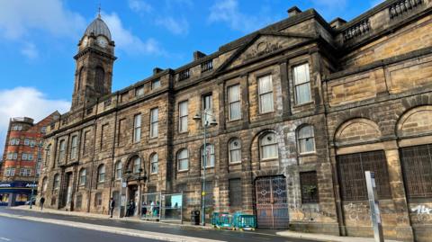 A three storey stone civic building with a clocktower. There is graffiti on the side of the building. A bus stop stands on the pavement outside the building and there is some green, red and white plastic safety barrier around part of the pavement.
