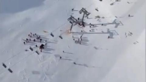 An aerial shot of a collapsed ski lift. A group of people can be seen gathered on the snow in the lower third of the shot.