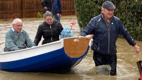 Simon O'Brien pulling people to safety in his homemade paddleboat. the road is flooded and an elderly man wearing a cardigan sits in the boat. A woman walks alongside.