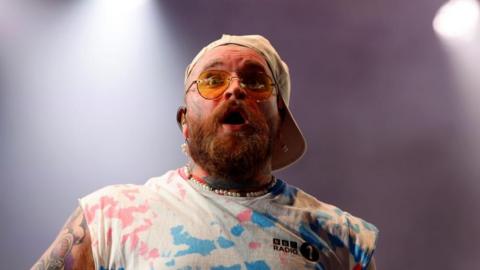 Singer Teddy Swims looking surprised - his mouth is open and his eyebrows are raised. He is wearing aviator-style sunglasses with yellow lenses and a baseball cap backwards. He has a ginger beard, and tattoos on his face and arms. He is wearing a white BBC Radio 1 top with blue and pink splashes and cut-off sleeves. 