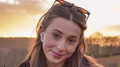 Darcy smiling at the camera. She has long brown hair and is standing in a field with the sun going down behind her. There is an orange glow around her. She has earpods in and sunglasses on her head.