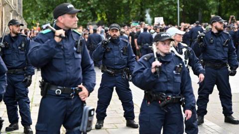 A group of five police officers in uniform holding batons on their shoulders and looking around at the crowds that have gathered for the protest in the city centre. 