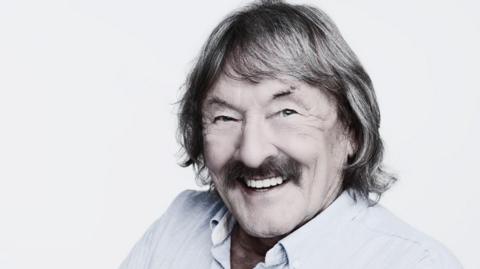 Trevor Campbell smiles to the camera. He has long shoulder-length brown hair and a brown moustache. He is wearing a pale white shirt.