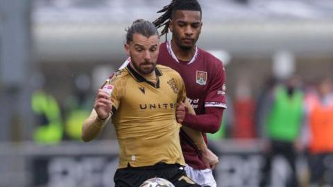 Jay Rodriguez in a gold Wrexham change shirt holds the ball off from a Northampton Town opponent