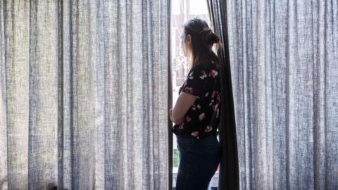 A stock image shows a woman looking out of a window in between partially drawn curtains with her arms folded.