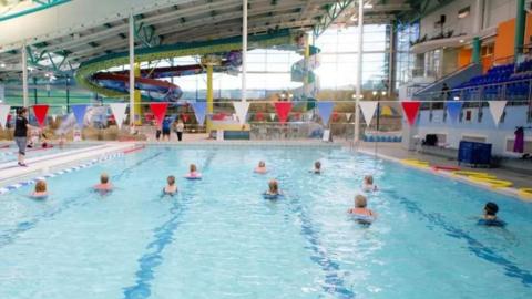 A large swimming pool at the NSC in Douglas with a group of people taking part in a class. There is a row of red, blue and white small triangular flags above the pool and water slides can be seen in the background.