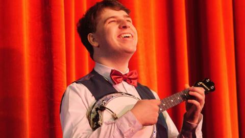 A close up shot of Matthew Quilliam playing his ukulele in front of some orange curtains wearing a red bow tie, brown waistcoast and pink shirt with the most enormous beaming smile on his face  