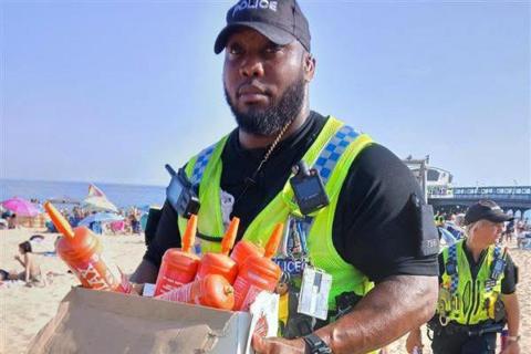 A male police officer wearing a black cap, short-sleeve t-shirt and a yellow hi-vis vest carries a box full of orange canisters on the busy sandy beach. Behind him is a female officer and Bournemouth Pier.