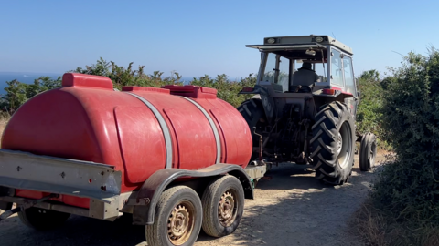 A huge red water container is being transported on the the back of a trailer pulled by a tractor on La Coupée linking Little Sark to Sark.