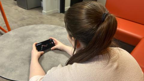An actress with her back to the camera poses with a smart phone which shows graphs detailing the current rate of cryptocurrency.