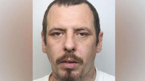 A police custody photo of Kieron Stuart Robert Long looking at the camera, with his mouth slightly open. He has short dark hair, a goatee beard and tattoos on his neck. He is wearing a white T-shirt.