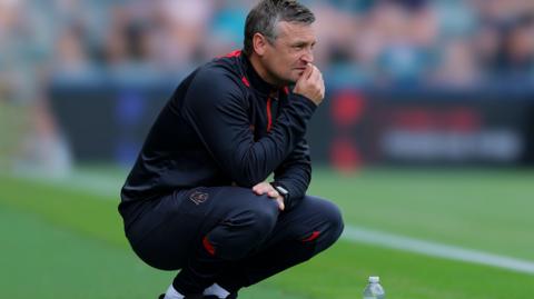 Bristol City manager Michael Skubala, in a dark blue tracksuit with red trim,  squats at pitchside, with his hand to his mouth watching his team in action.