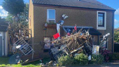 A semi-detached house with wood and other household items cluttered in the front garden along the side. 