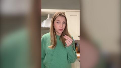 A woman in a green jumper stands in a kitchen