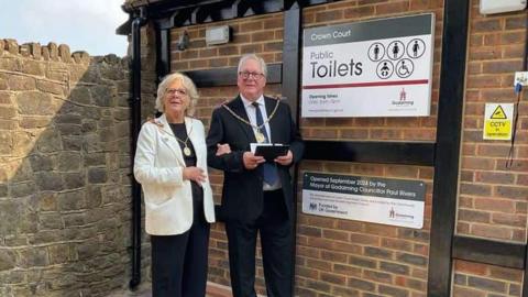 The Mayor and Mayor Consort dressed in their ceremonial chains stand outside the toilets made out of red bricks. A plaque on the toilet wall reads "Opened September 2024 by the Mayor of Godalming councillor Paul Rivers"