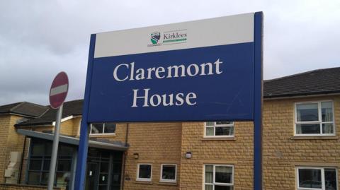 A two-storey building with sand-coloured bricks and windows with white frames. In the foreground is a blue sign saying 'Claremont House'. 