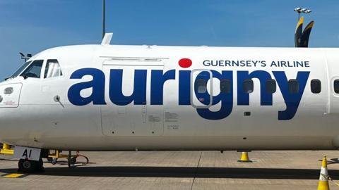 A picture of a white plane with the Aurigny branding on the side of it. It is parked at an airport. The logo is blue and red in colour. There are yellow cones around it