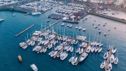 The image shows an aerial view of a marina, with many sailing boats and yachts docked at multiple pontoons. A breakwater surrounds the marina, and buildings line the shore. 