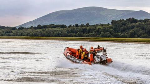 A lifeboat skipping across the water surface on the Solway estuary with trees and hills in the background