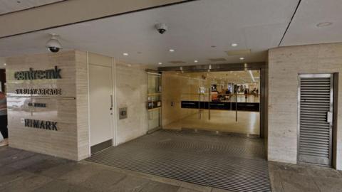 An entrance into a shopping centre, which is under cover. There are four glass doors with silver handles, and through them is a lit corridor. Signs on the outside wall, which is pale marble-effect, say "centre:mk", "Silbury Arcade" and "Primark".