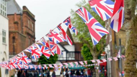 Union Jack bunting is hung up in a street. There are trees and buildings in the background and people walking around