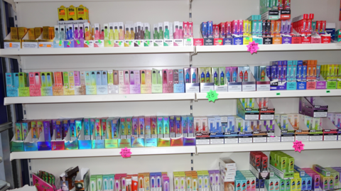 Rows of vapes are on sale in the shop. Four shelves of goods are visible in the image, all different colours with price tags also on display.