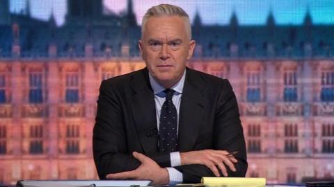 Huw Edwards, who has grey hair, wearing a dark jacket and blue tie over a light shirt