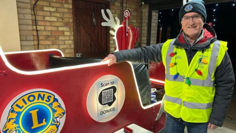 A man wearing glasses, a dark beanie hat and a hi-vis tabard over a dark coat and jeans with Christmas lights hanging around his neck stands next to a wooden sleigh. The sleigh is red and decorated with the insignia of Lions Clubs International and a QR code labelled "Scan to donate".
