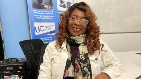 A woman with wavy hair. She is wearing a white jacket and multi-coloured shirt. She is sitting by a desk.