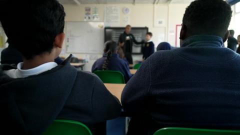 The back of two pupils sitting in plastic chairs as they watch a role play at the front of the class