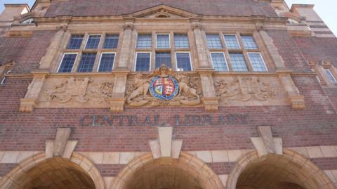 The main entrance of Wolverhampton Central Library seen from street level, looking up at the ornate first-floor façade. A colourful royal coat of arms sits centrally above the 'Central Library' lettering, flanked by carved stone lions. 