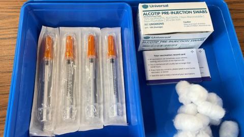 Vaccination syringes on a blue tray. A box of wipes and some cotton wool are also on the tray.