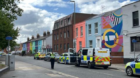 Three police cars and a police van park alongside colourful houses on a cordoned off road in Gloucester city centre. A police officer can be seen standing behind the police tape while another, who is on the phone, gets out of the van.
