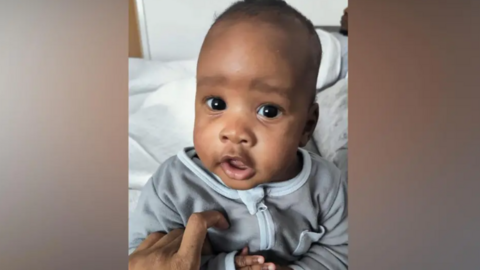 Noah is in a grey baby-suit. He is looking at the camera and appears to be sat on on a bed in a bedroom.