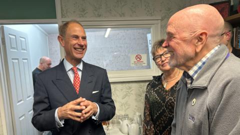 Duke of Edinburgh wearing a suit, blue shirt and red tie smiling at a man dressed in a grey zip-up top. 