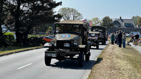 Convoy of vintage military trucks driving down a suburban road, with spectators watching from the roadside. The lead truck, marked with a star emblem and license plate 'YXS 966', is followed by similar vehicles under clear skies.