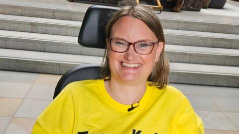 Kate wearing a bright yellow T-shirt smiling at the camera. She is wearing glasses and has mid-length brown hair. She is sitting in her wheelchair in front of a set of steps.